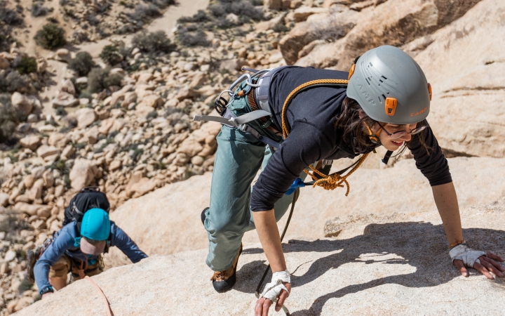 A person wearing safety gear climbs a rock incline, with another person below them.